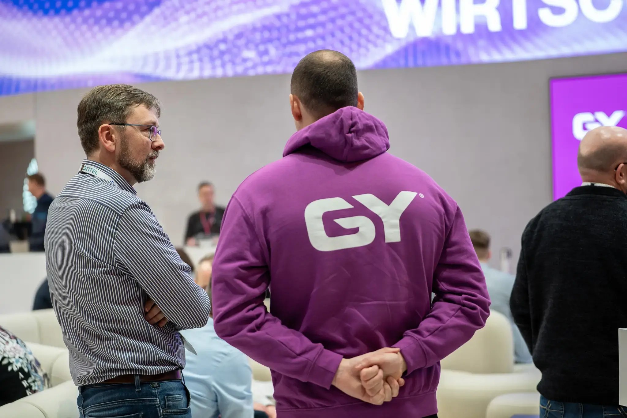 Zwei Männer wurden von hinten fotografiert. Dabei trägt einer von ihnen einen lila Hoodie mit einem weißen Logo auf dem Rücken, bestehend aus den Buchstaben G und Y. Es scheint, dass sich die beiden Männer unterhalten.