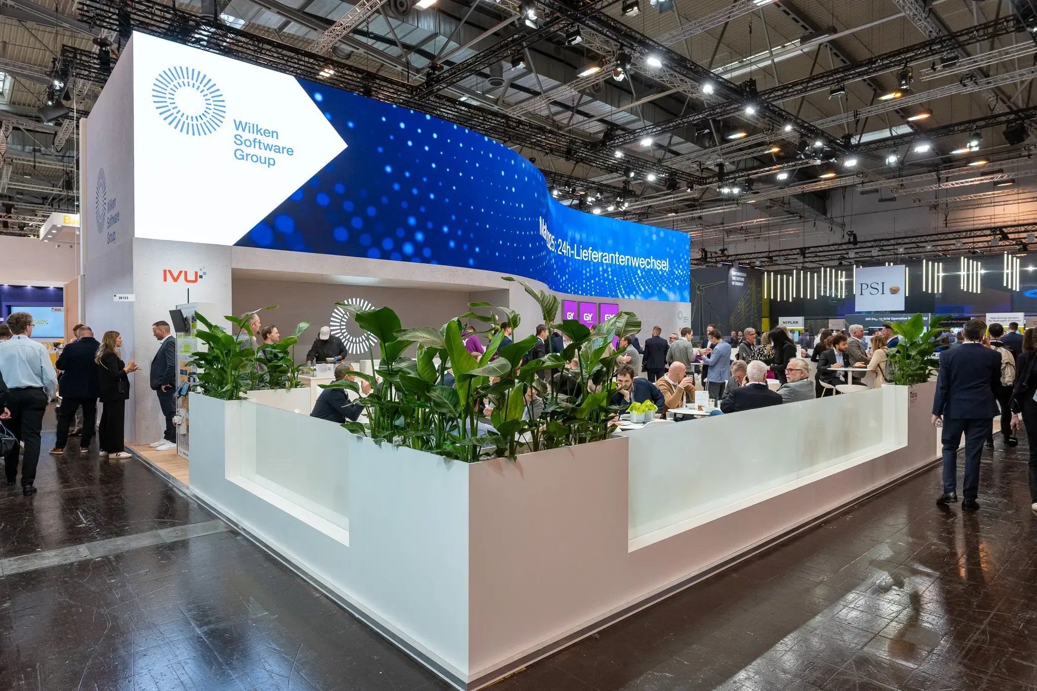 Eine Gesamtaufnahme eines sehr offenen Messestandes der Wilken Software Group. Auffällig ist eine große, wellenförmige LED-Wand, auf der verschiedene Botschaften gespielt werden. Im Vordergrund des Messestandes ist eine grüne und durch weiße Abgrenzung, Sitzecke für Gespräche zu sehen.