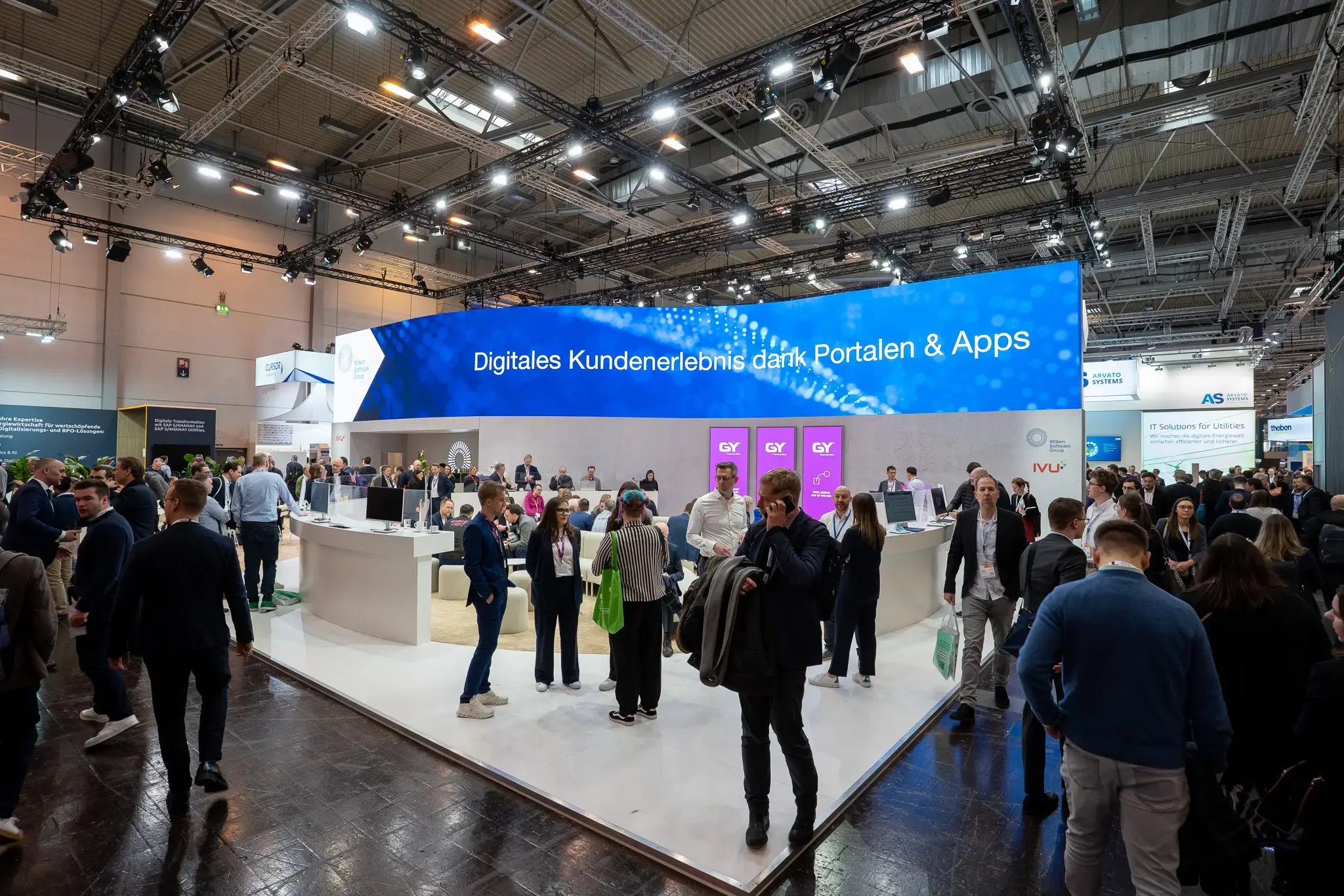 Ein großer, offener Messestand der von vielen Menschen gut besucht ist. Es gibt viele Gespräche und einige Besucher laufen am Stand vorbei. Auffällig ist, dass der Stand eine große blaue LED-Wand oberhalb der Rückwand hat, mit der Botschaft: Digitales Kundenerlebnis dank Portale & Apps.