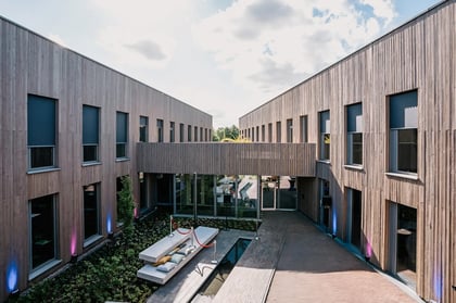 Moderner Eingangsbereich eines Bürogebäudes mit Holzfassade, großen Fensterfronten und begrüntem Innenhof. In der Mitte Sitzgelegenheit mit Polstern über einem Wasserlauf. Brückenverbindung im ersten Stock und dezente Beleuchtung an der Fassade.