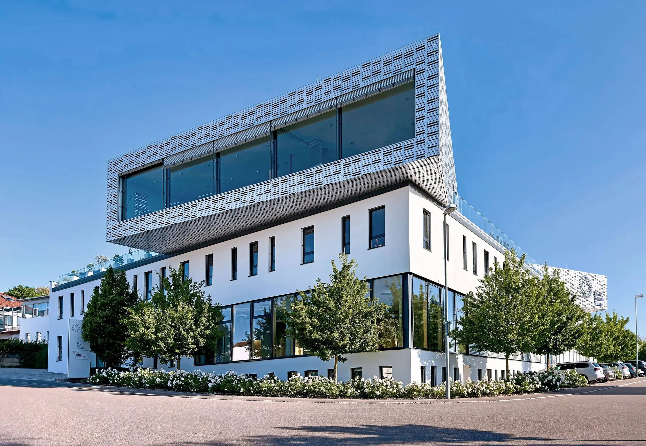 Modernes Firmengebäude der Wilken Software Group in Ulm mit markanter Architektur: Weiße Fassade, große Glasflächen und ein gläserner Aufbau, der über das Hauptgebäude hinausragt. Umgeben von Bäumen, Blumenbeeten und einer sonnigen Umgebung.