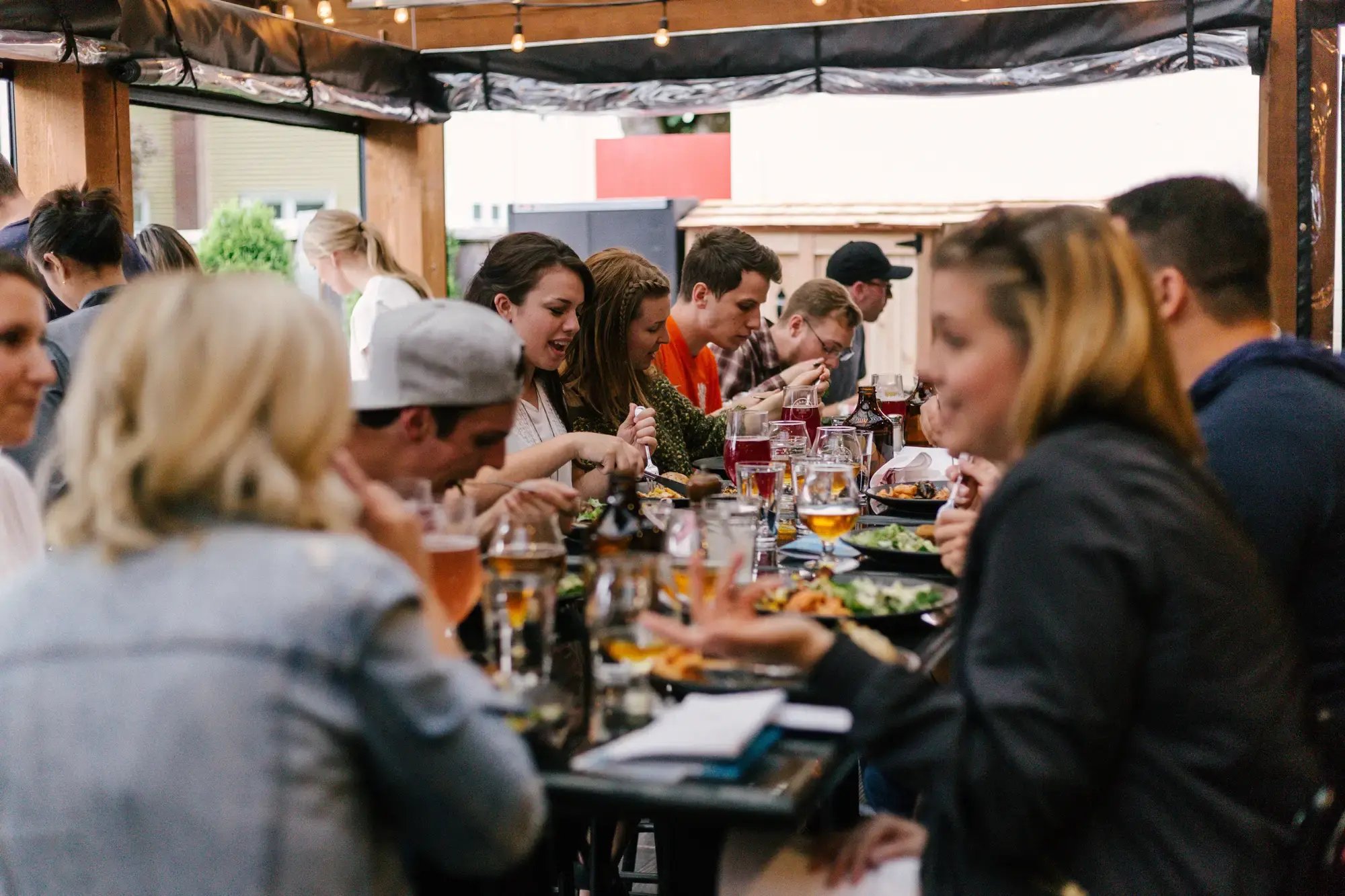 Mehr als fünf Personen sitzen gemeinsam beim Essen und Trinken an einem Tisch und unterhalten sich während dem Essen. Es scheint, als ob die Personen an einem Sommermittag oder Abend draußen sitzen.