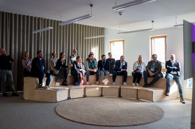 Eine Gruppe von Menschen sitz auf einer Holzarena bzw. Podest und schauen interessiert in einen Bildschirm, auf dem etwas abgespielt wird.