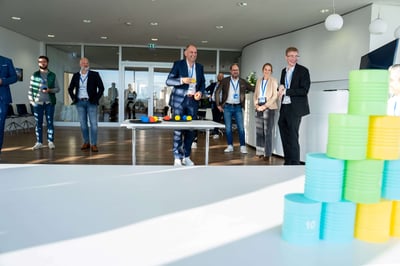 Geschäftsleute spielen in einer Gruppe Dosenwerfen. Eine Person wirft und die anderen schauen zu. Die Stimmung wirkt locker und alle haben Spaß.