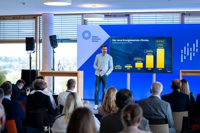 Ein Mann steht auf einer Bühne und präsentiert seine Folien vor einer größeren Gruppe von Menschen, die vor ihm sitzen. Dabei handelt es sich um eine kleinere Tagung.