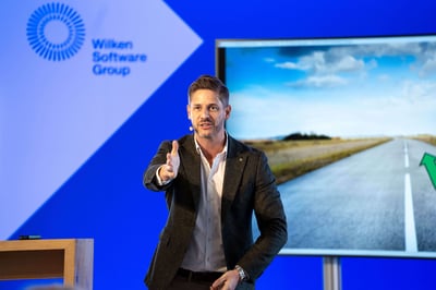 Ein Mann mittleren Alters steht auf einer Bühne und vor einem Bildschirm und präsentiert. Dabei richtet er das Wort gezielt an die Teilnehmer, da seine Hand in die Richtung des Bildes geht. Der Speaker trägt dabei sein Mikro an sich.