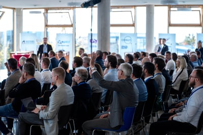 Eine Gruppe von Geschäftsleuten sitzen in einem hellen Raum und hören einem Vortrag zu. Dabei ist der Präsentierende nicht zu sehen, aber es gibt aus dem Publikum eine Wortmeldung eines Mannes.