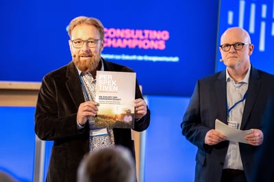 Zwei Männer mit Mikros stehen auf einer Bühne. Einer der Männer hält ein Magazin in der Hand und präsentiert dieses den Teilnehmern vor ihm. Der andere Mann steht mit seinen Morderationskarten neben ihm.