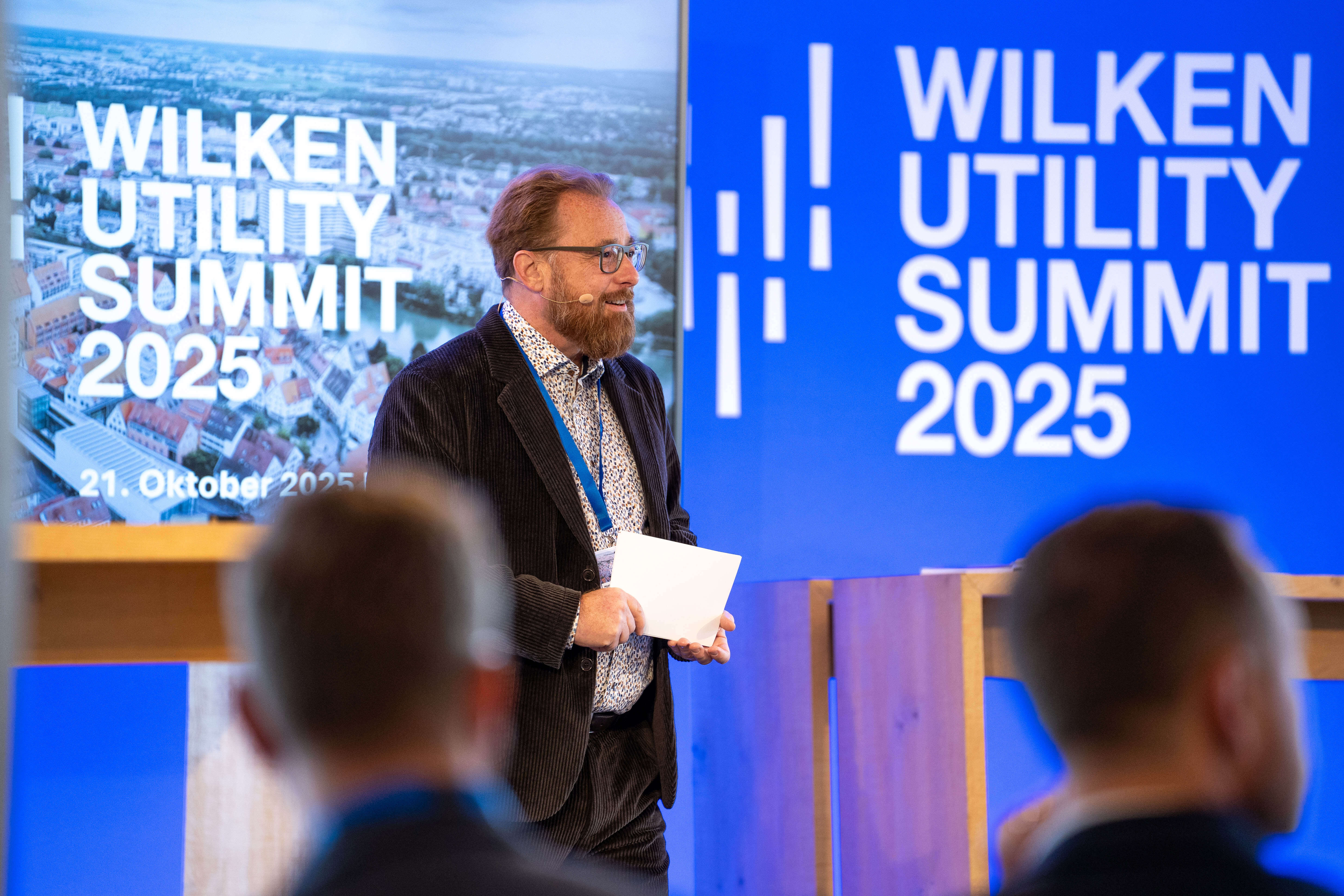 Ein Mann mit Brille und Headset-Mikrofon steht auf einer Bühne vor Teilnehmern des Wilken Utility Summits 2025.