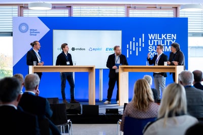 Fünf Männer und Partner der Wilken Software Group stehen auf einer Bühne des Wilken Utility Summits 2025. Auf der Bühne findet eine Diskussion unter den Personen statt, die durch einen Moderator in der Bitte geführt wird. Die Teilnehmer stehen hinter etwas höheren Holztischen.