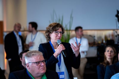 Eine Frau steht in der Mitte von sitzenden Teilnehmern und hält ein Mikrofon in der Hand, um etwas zu sagen. Es handelt sich dabei um eine Businessveranstaltung.