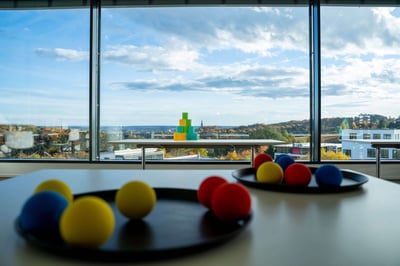 Ein Tisch mit zwei Tabletts, auf denen bunte Bälle liegen, die zum Dosen werfen vorgesehen sind. Im Hintergrund sind die bunten Dosen zu sehen, sowie eine große Fensterfront. Aus dem Fenster heraus sieht man einen herbstlichen Tag mit Sonne.