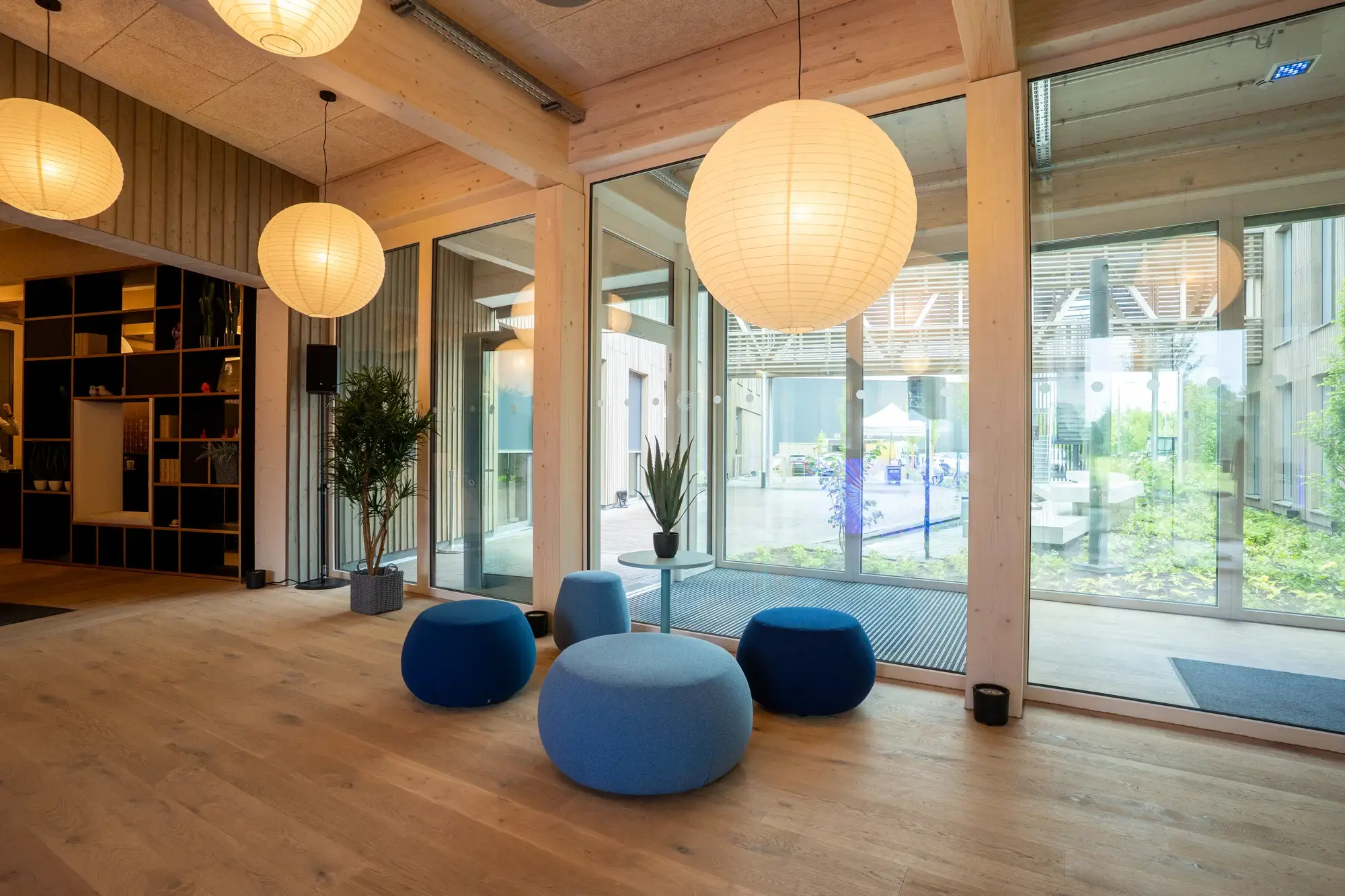 Moderner Empfangsbereich im Wilken-Gebäude in Greven mit großen Fenstern, hölzernem Interieur, runden Hängelampen und einer gemütlichen Sitzgruppe aus blauen Hockern und einem kleinen Tisch mit Pflanze – mit Blick auf den begrünten Innenhof.