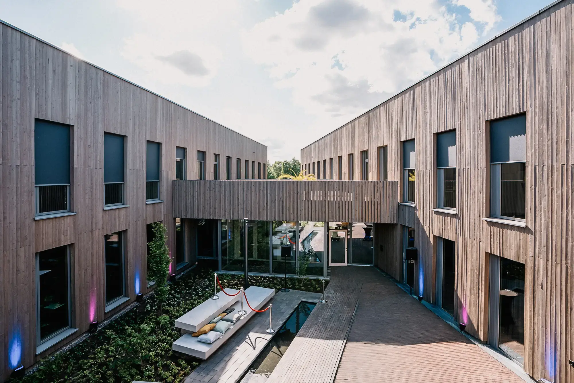 Moderner Eingangsbereich eines Bürogebäudes mit Holzfassade, großen Fensterfronten und begrüntem Innenhof. In der Mitte Sitzgelegenheit mit Polstern über einem Wasserlauf. Brückenverbindung im ersten Stock und dezente Beleuchtung an der Fassade.