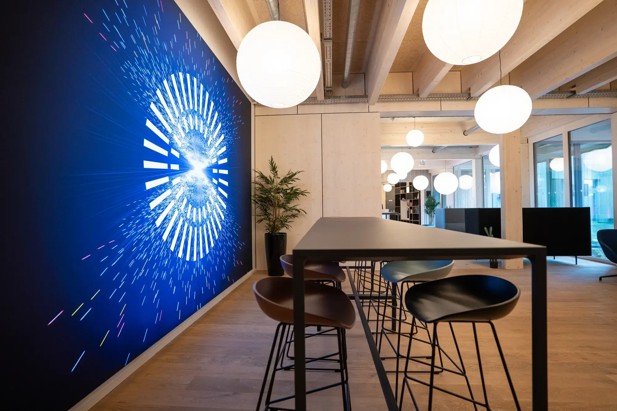 Moderner Open Space im Wilken-Gebäude in Greven mit hoher Decke, runden Hängelampen, Hochtisch mit Barhockern und leuchtender, digital wirkender Grafik an der Wand – stilvolles Ambiente für kreativen Austausch.