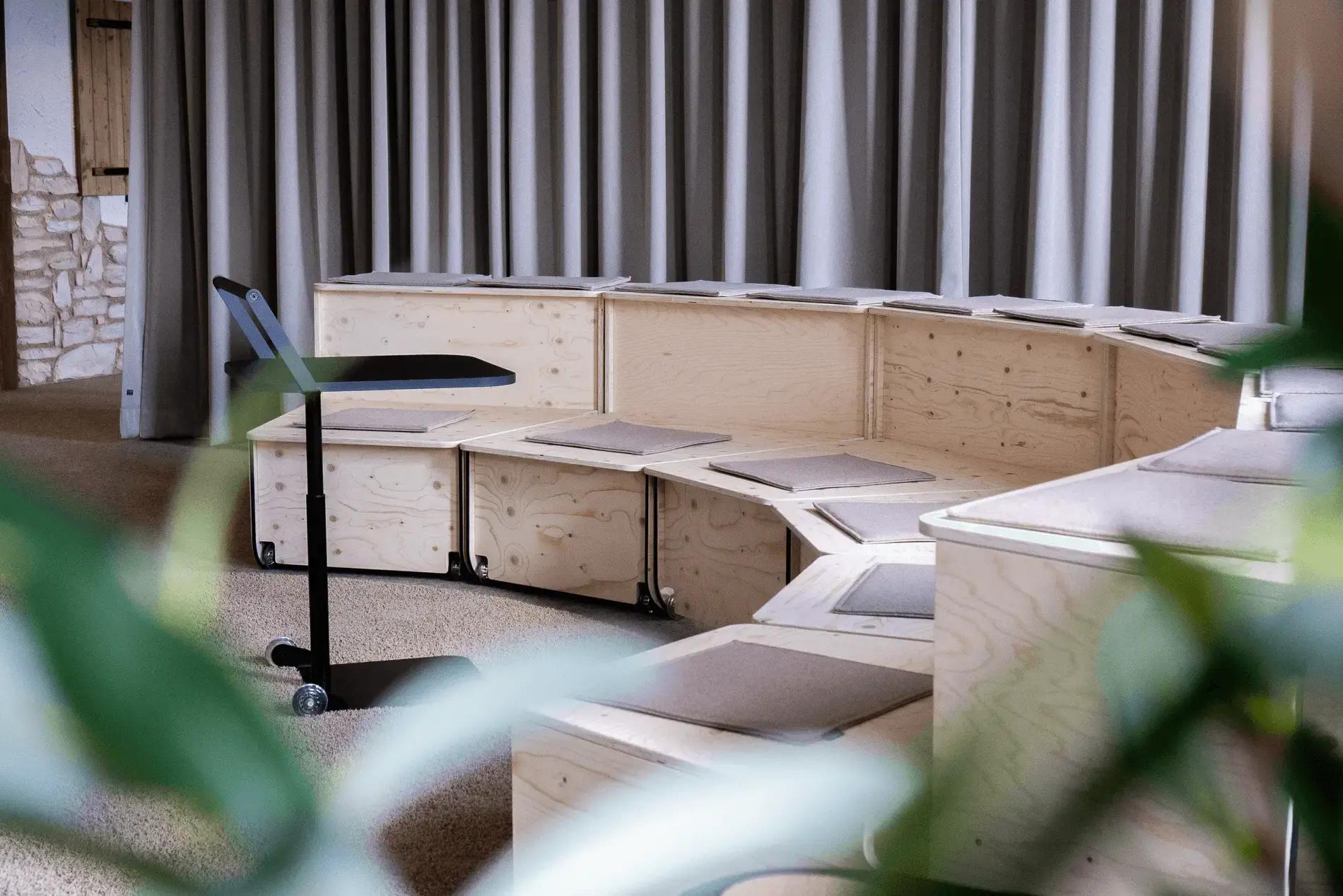 Moderne amphitheaterartige Sitzreihen aus Holz sind zu sehen. Braune dünne Kissen dienen als Sitzunterlage und lassen die Sitzmöglichkeiten gemütlicher wirken. Das Bild wurde durch eine Pflanze fotografiert, wodurch im Vordergrund grüne unscharfe Blätter zu erkennen sind.