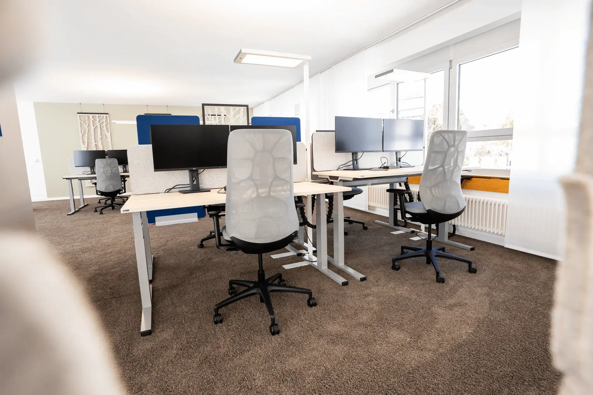 Ansicht eines modernen Großraumbüros mit Fokus auf Komfort und Funktionalität. Ein brauner Hochfloorteppich sorgt für eine angenehme Atmosphäre, während helle Tische und Stühle mit Bildschirmen eine produktive Arbeitsumgebung unterstützen.