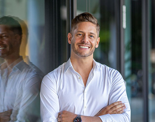Portraitbild eines jungen Mannes in einem weißen Hemd vor einem Gebäude mit vielen Glasscheiben. An der linken Hand trägt er eine Uhr. Portraitbild eines jungen Mannes in einem weißen Hemd vor einem Gebäude mit vielen Glasscheiben. An der linken Hand trägt er eine Uhr.